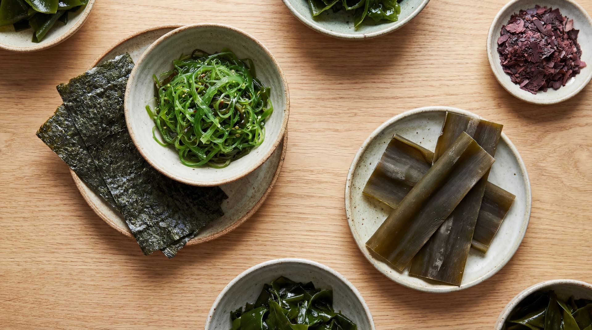 Assorted edible seaweeds in ceramic bowls on a wooden table, showing nori, wakame, kombu, and dulse.