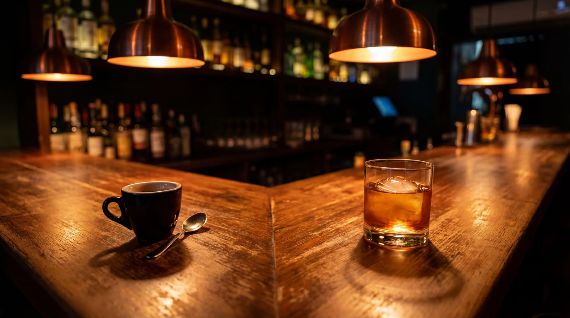 Dark espresso cup and amber cocktail glass on a polished wood bar counter under warm moody lighting
