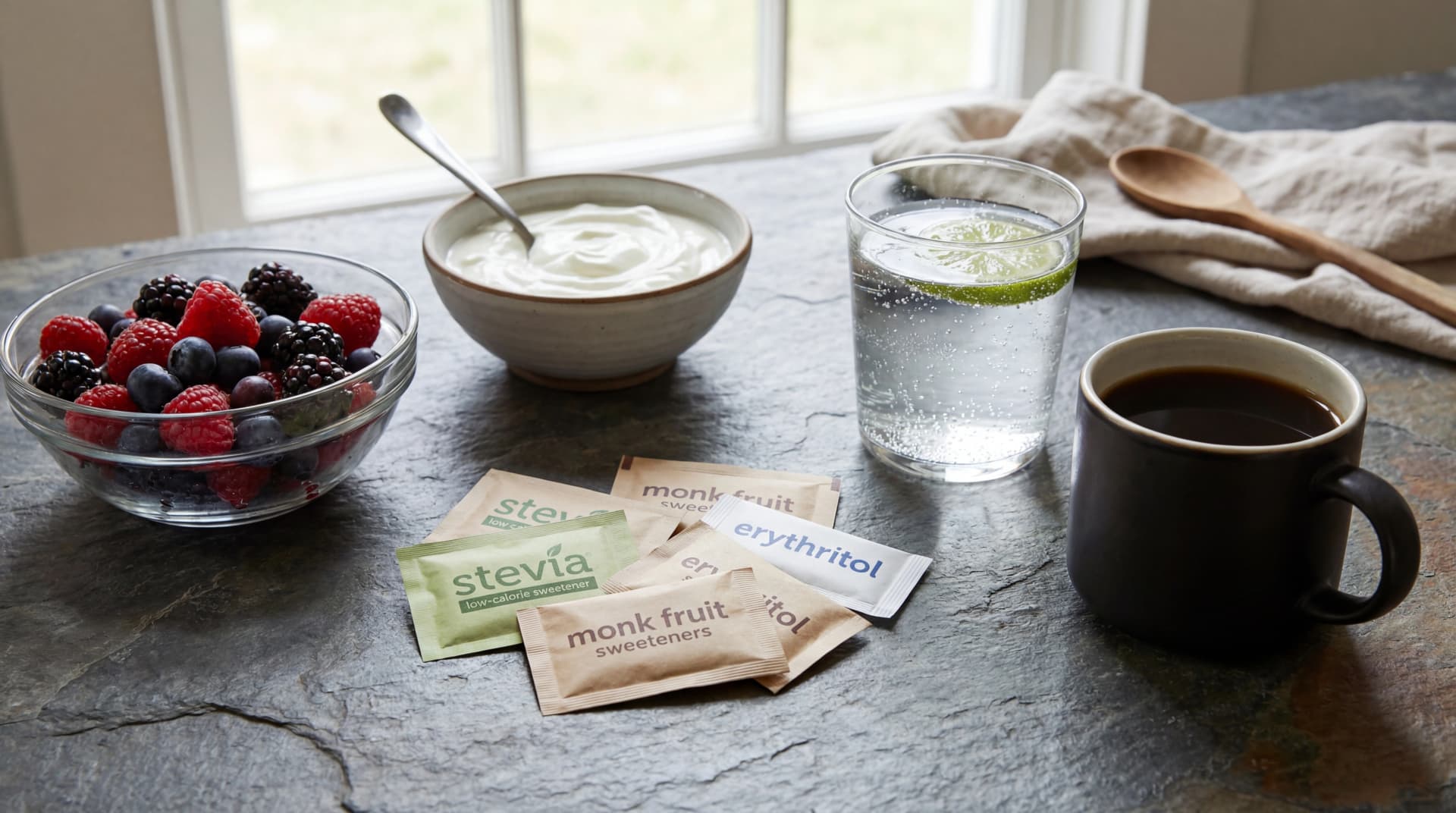 Assorted low-calorie sweetener packets with berries, yogurt, sparkling water, and black coffee on a kitchen countertop.